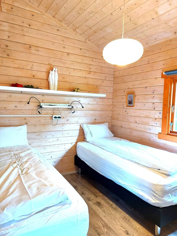 A wooden cabin room with two beds, a shelf with decorative items, and a hanging light fixture. - Ásgeirsstaðir Cottages (Eidar)