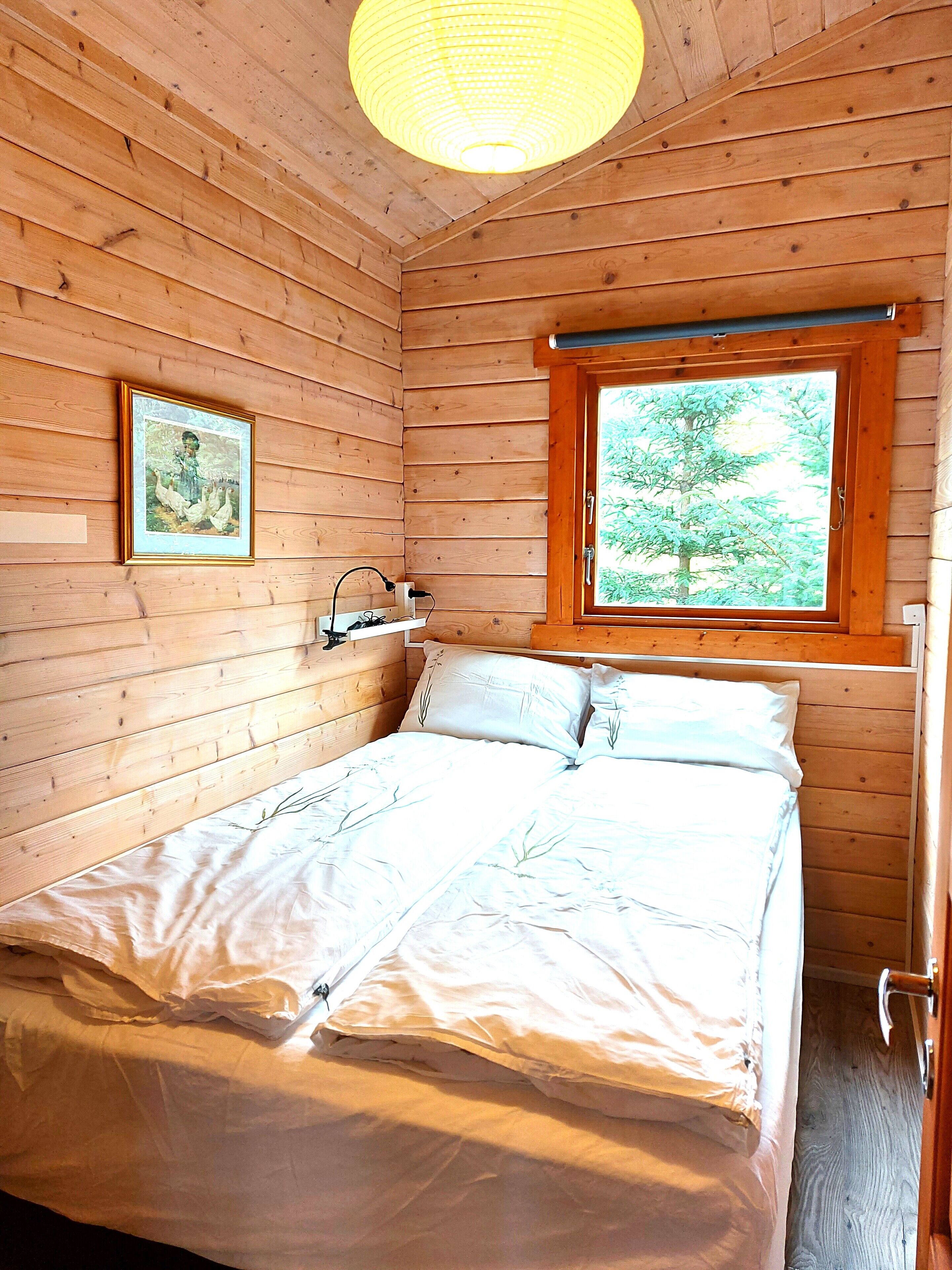 A wooden room with a bed, a window with a view of trees, and a framed picture on the wall.