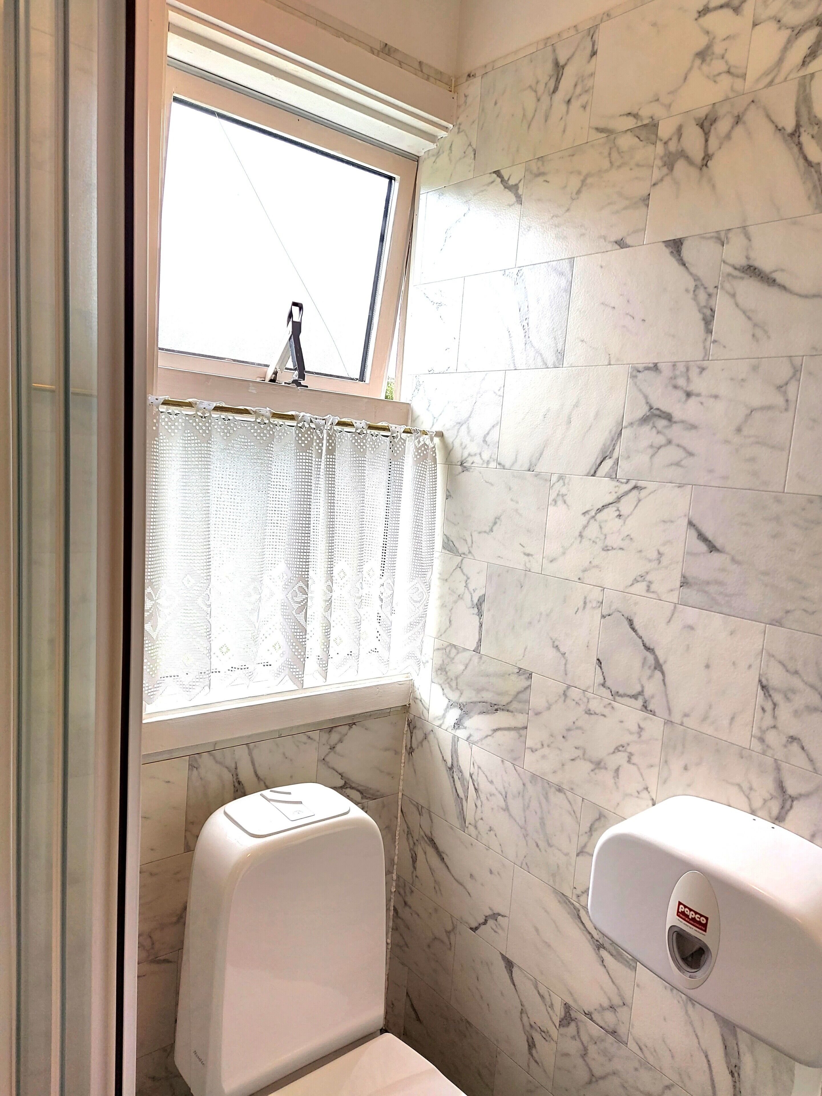 A bathroom with a marble wall, a window with a lace curtain, a toilet, and a hand dryer.