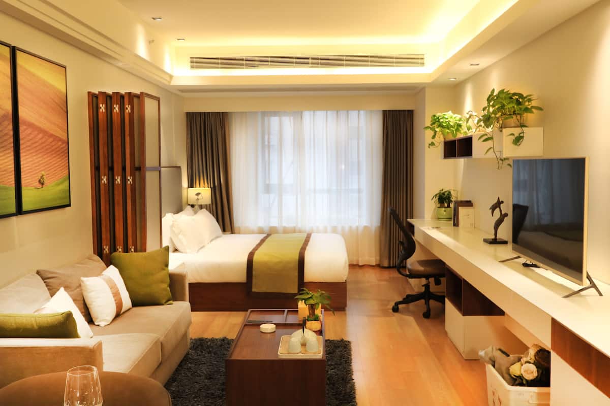 A modern hotel room with a bed, sofa, desk, TV, and potted plants.