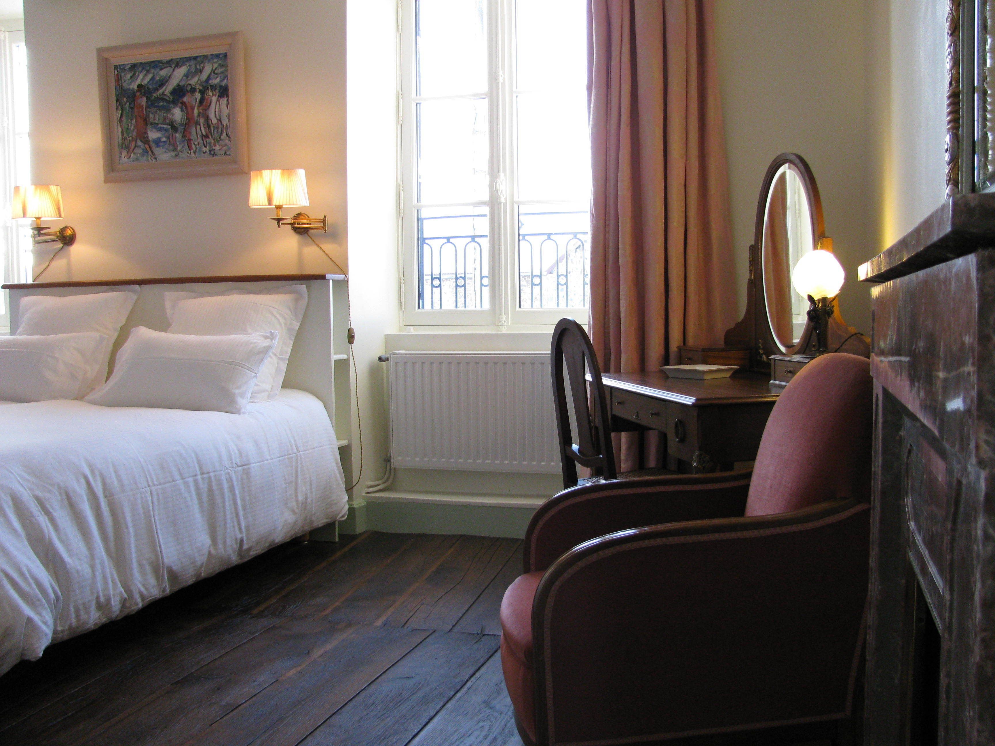 A hotel room with a bed, a desk with a chair, a dresser, a window with curtains, a radiator, and a painting on the wall.