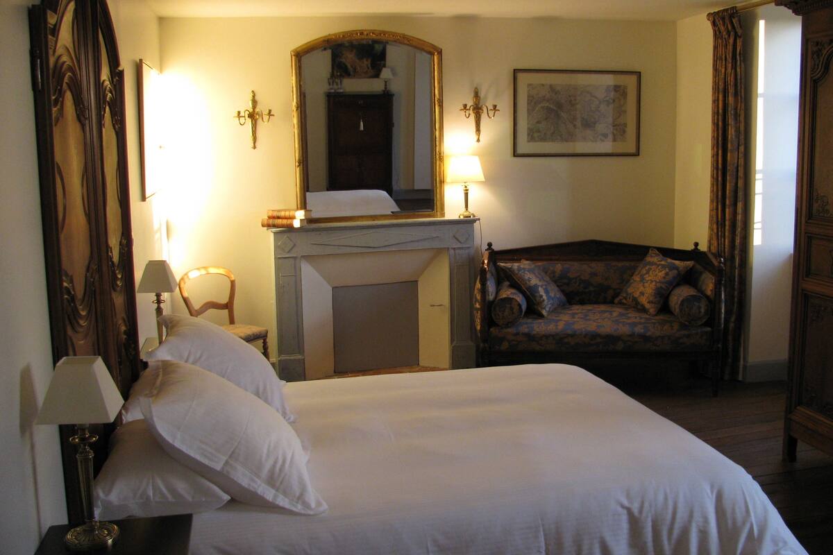 A hotel room with a large bed, a chair, a fireplace, and a mirror.