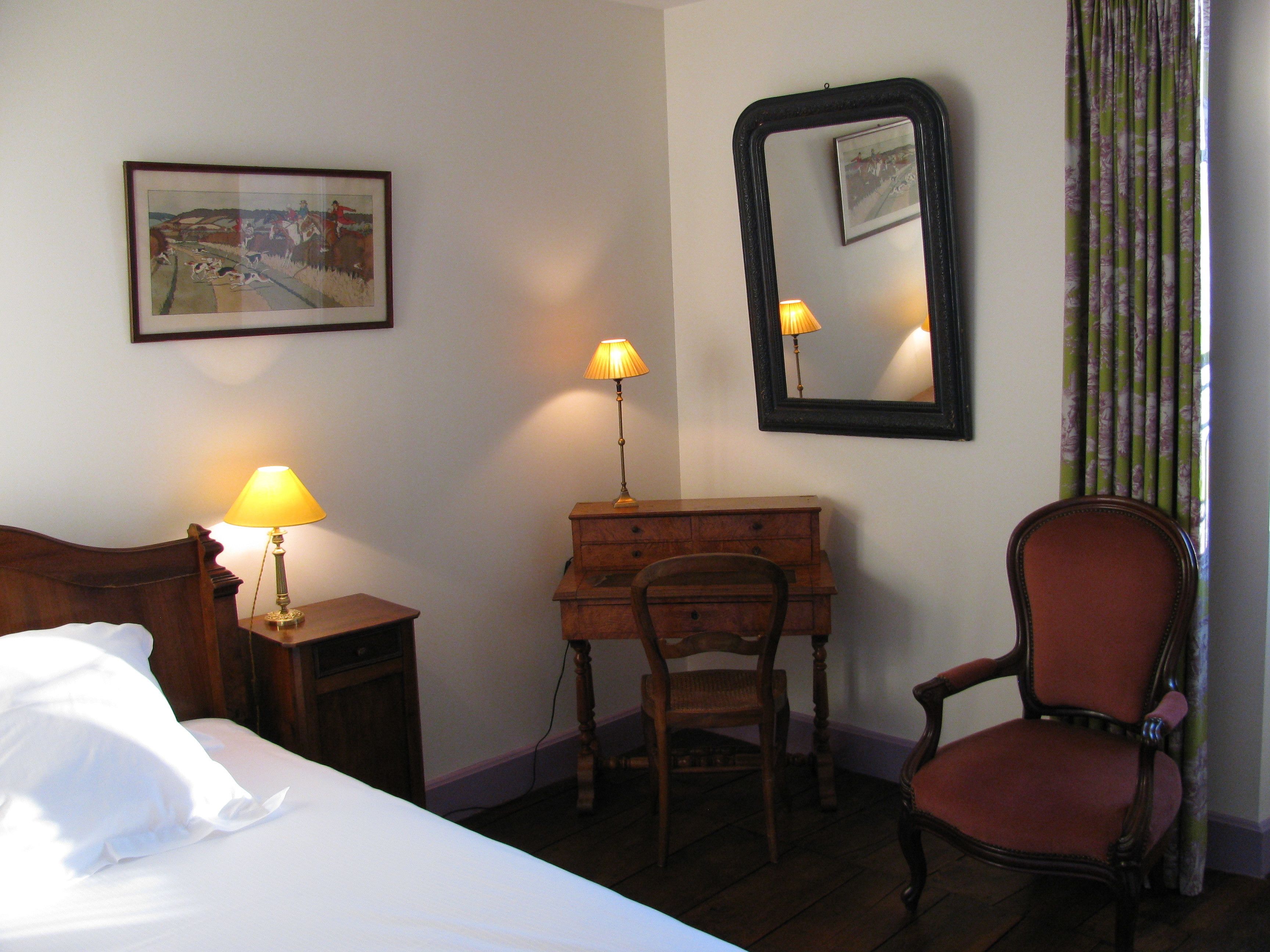 Habitación de hotel con cama, un escritorio de madera, una silla, cómoda, lámpara, espejo y un cuadro en la pared.