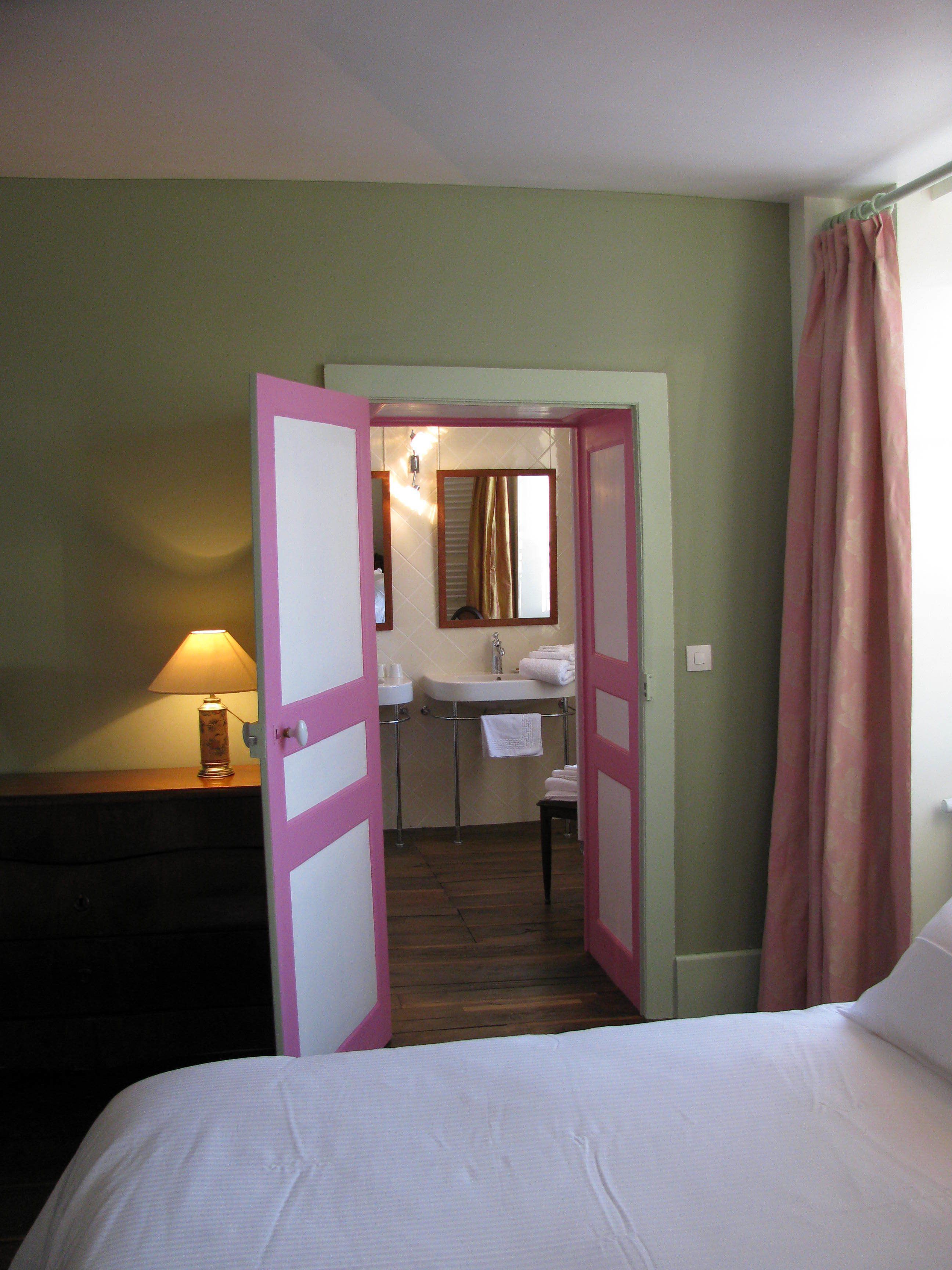 Hotelkamer met een bed, een nachtkastje met een lamp, een badkamer met een wastafel en spiegel, en een deur die naar een andere kamer leidt.