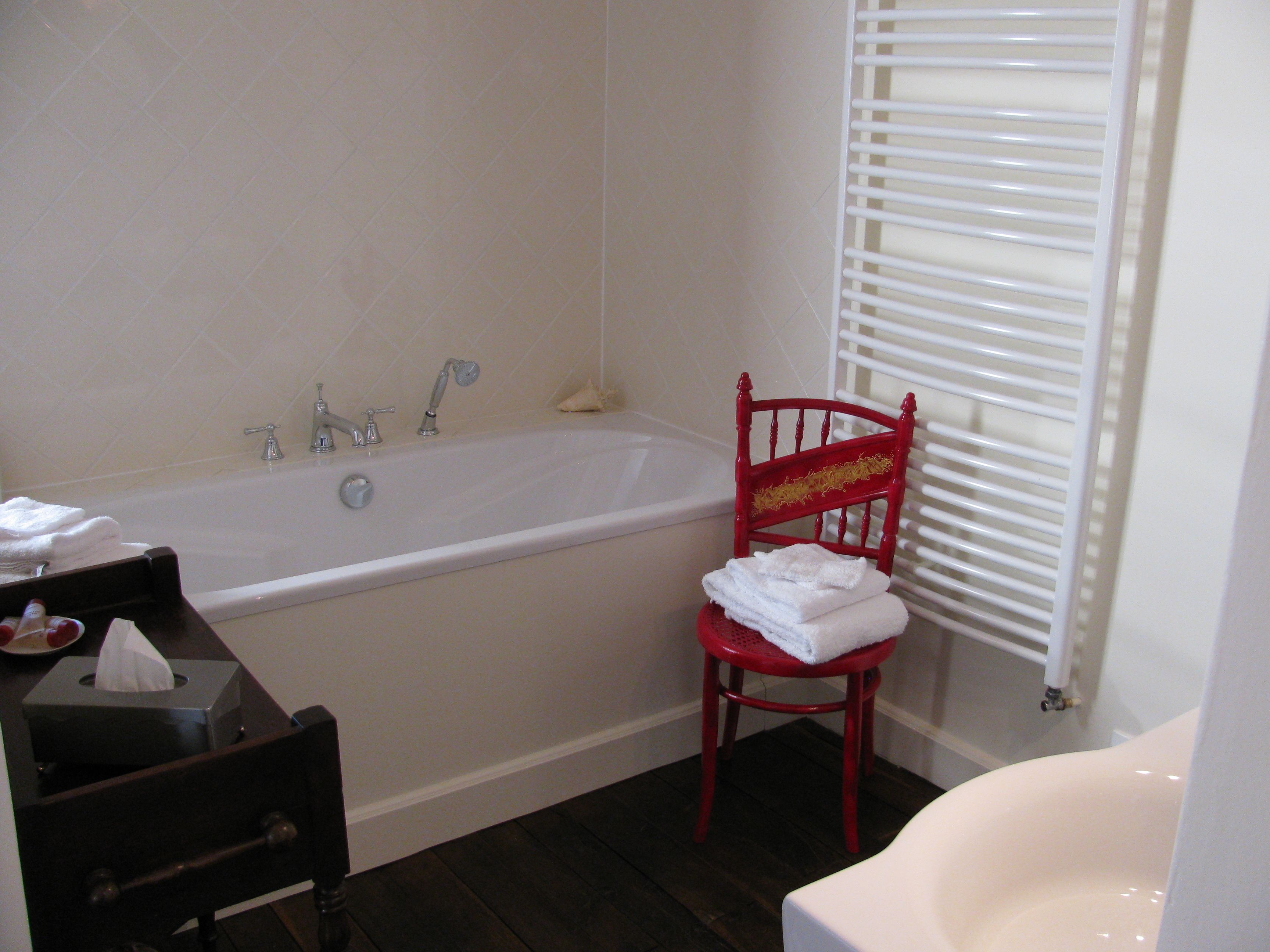 Un baño con una bañera blanca, una silla roja con toallas, suelo de madera oscura y un inodoro blanco.