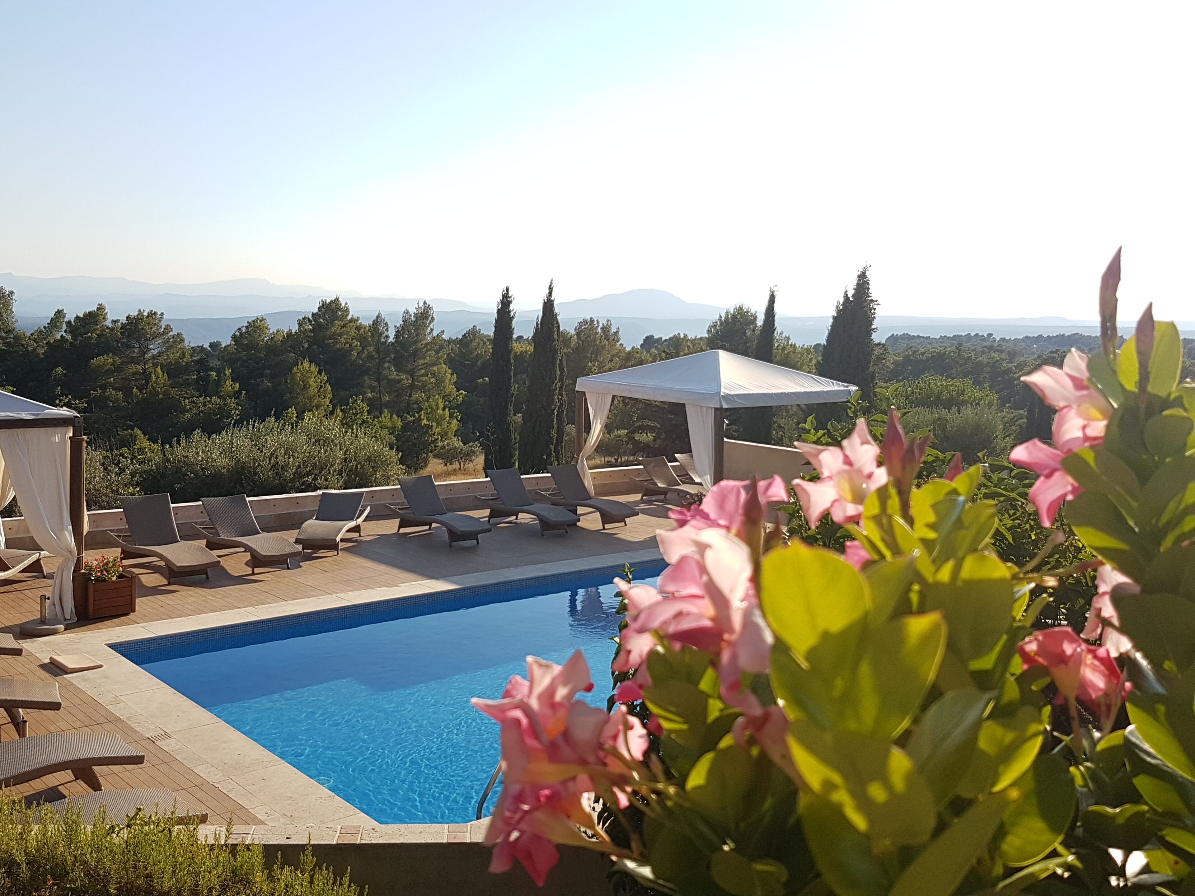 A pool area with lounge chairs and a pergola, surrounded by greenery and a view of distant mountains.