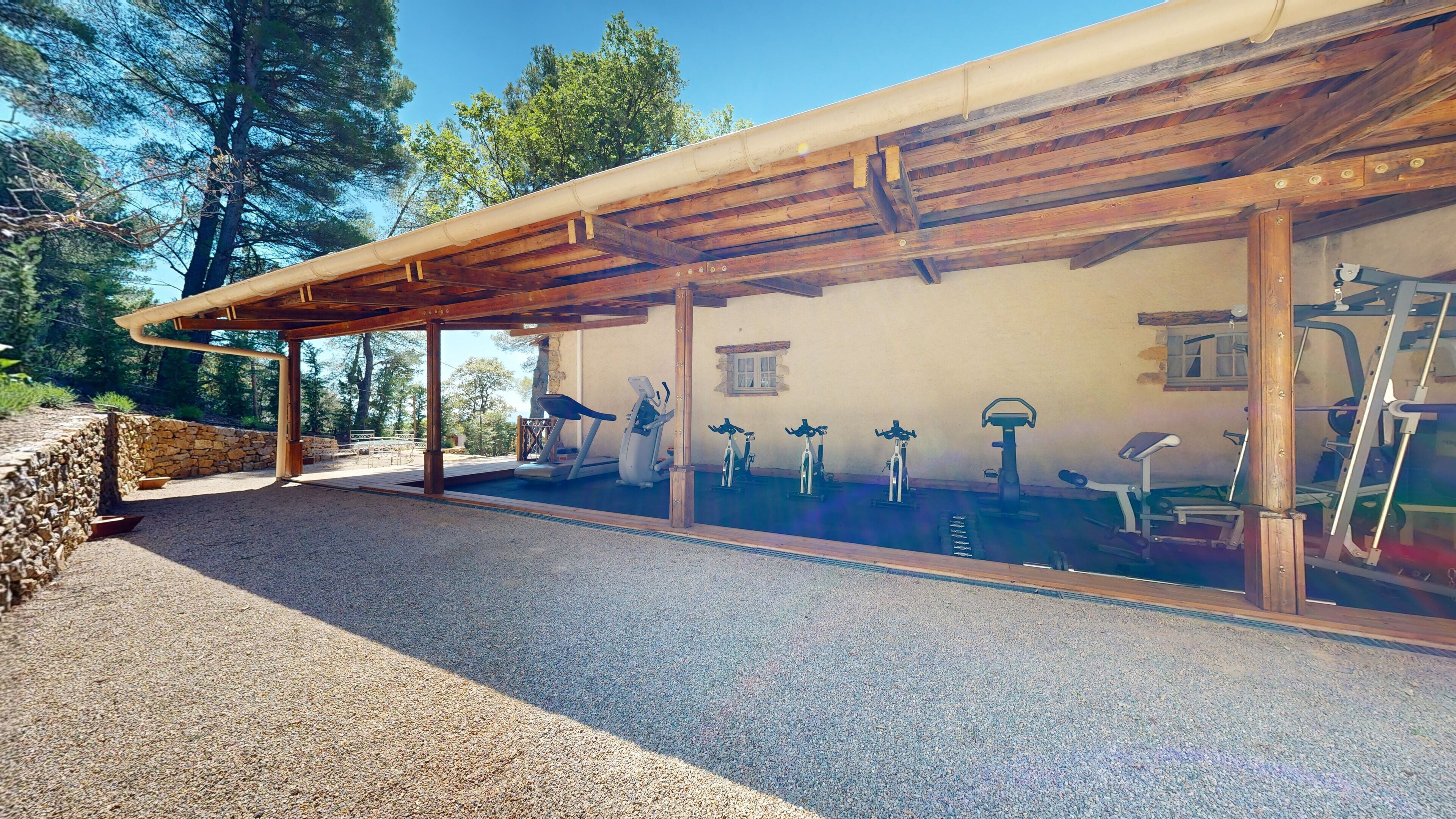 Un espace de remise en forme extérieur abrité, équipé de matériel d’exercice, avec un mur en pierre et une vue sur les arbres.