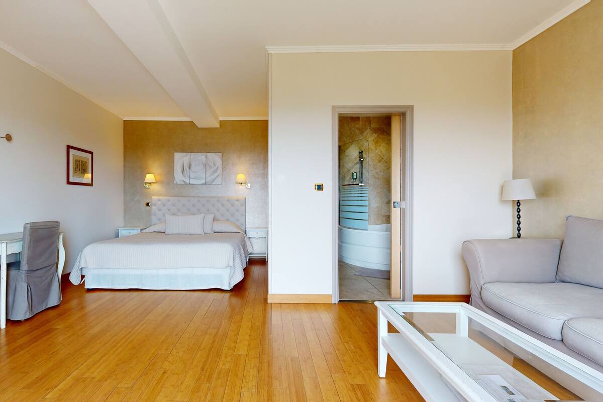 A hotel room with a bed, a desk, a chair, a sofa, and a bathroom visible through an open door.