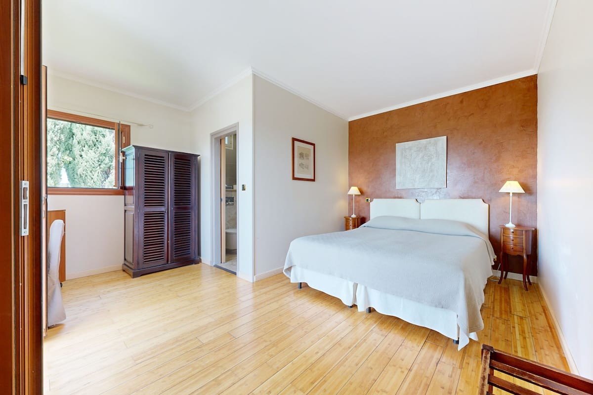 A bedroom with a wooden floor, a bed with a white bedspread, two bedside tables with lamps, a wardrobe, and a framed picture on the wall.