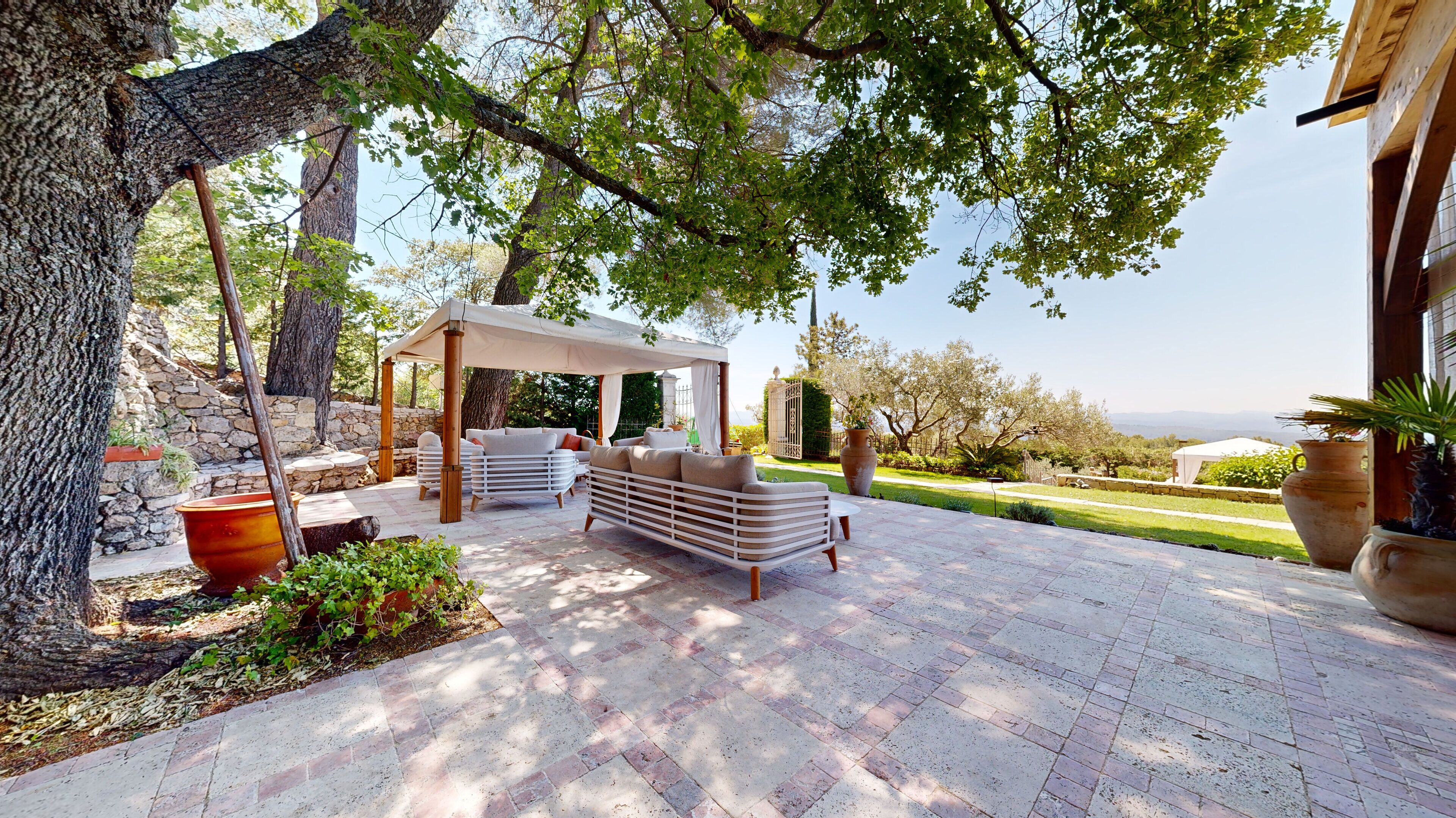 A covered outdoor seating area with a sofa and chairs, surrounded by trees and a stone wall.