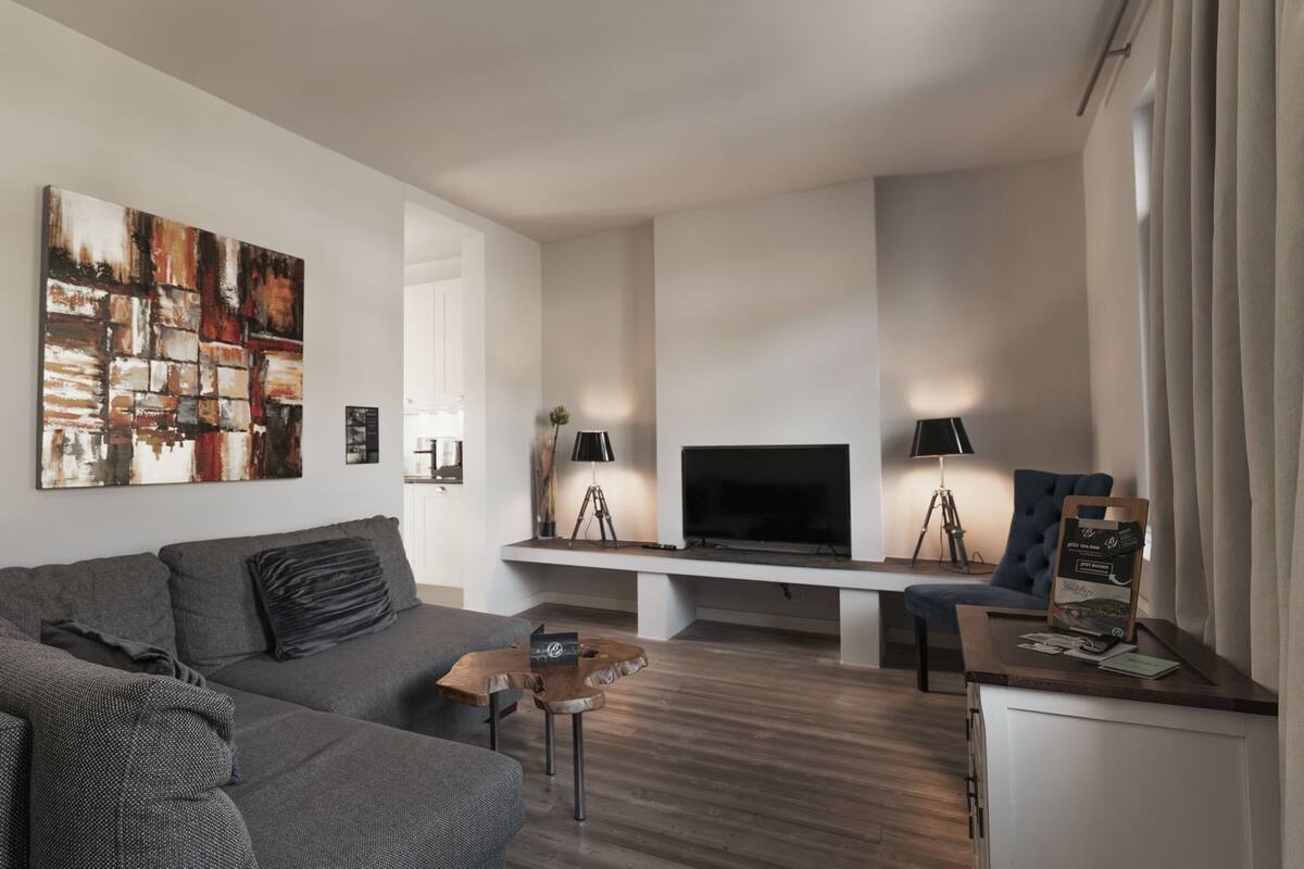 Ein modernes Wohnzimmer mit grauem Sofa, einem Holztisch, einem Fernseher auf einem Schrank und einem Schreibtisch mit Lampe.