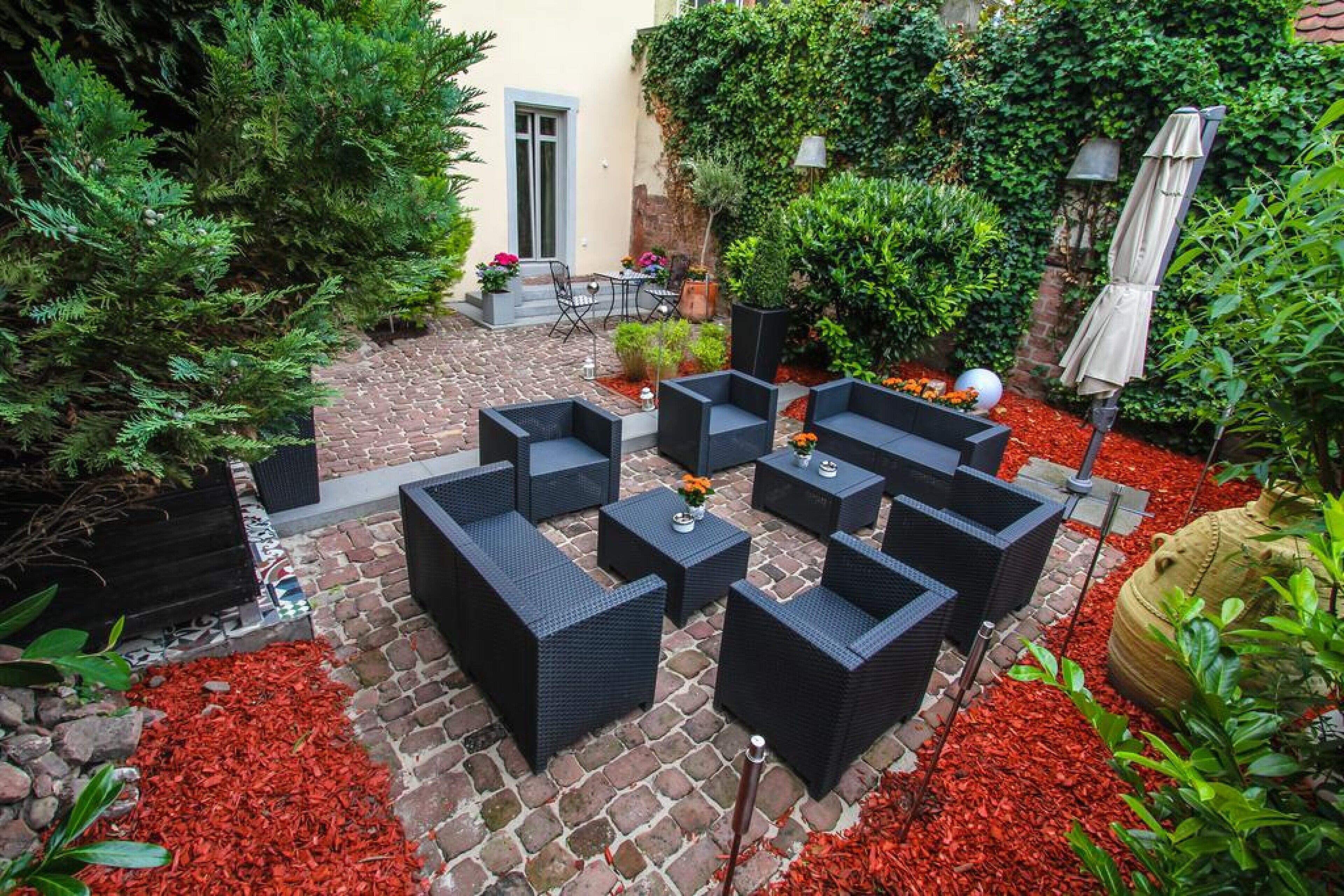 Un patio al aire libre con mobiliario de mimbre negro, cobertura de mantillo rojo y diversas plantas.