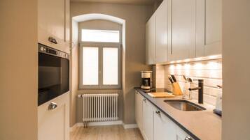 A compact kitchen with a built-in oven, a window with blinds, a radiator, and a countertop with a coffee maker and utensils.