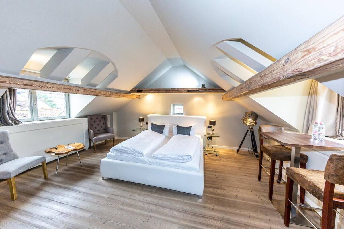 Ein Schlafzimmer mit einem Bett, zwei Stühlen, einem kleinen Tisch und einem Schreibtisch mit Stühlen. Es gibt Dachfenster und ein Fenster.