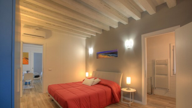 A bedroom with a bed, bedside lamps, a painting on the wall, and a bathroom with a towel rack. - Ca Sarasina (Venice)