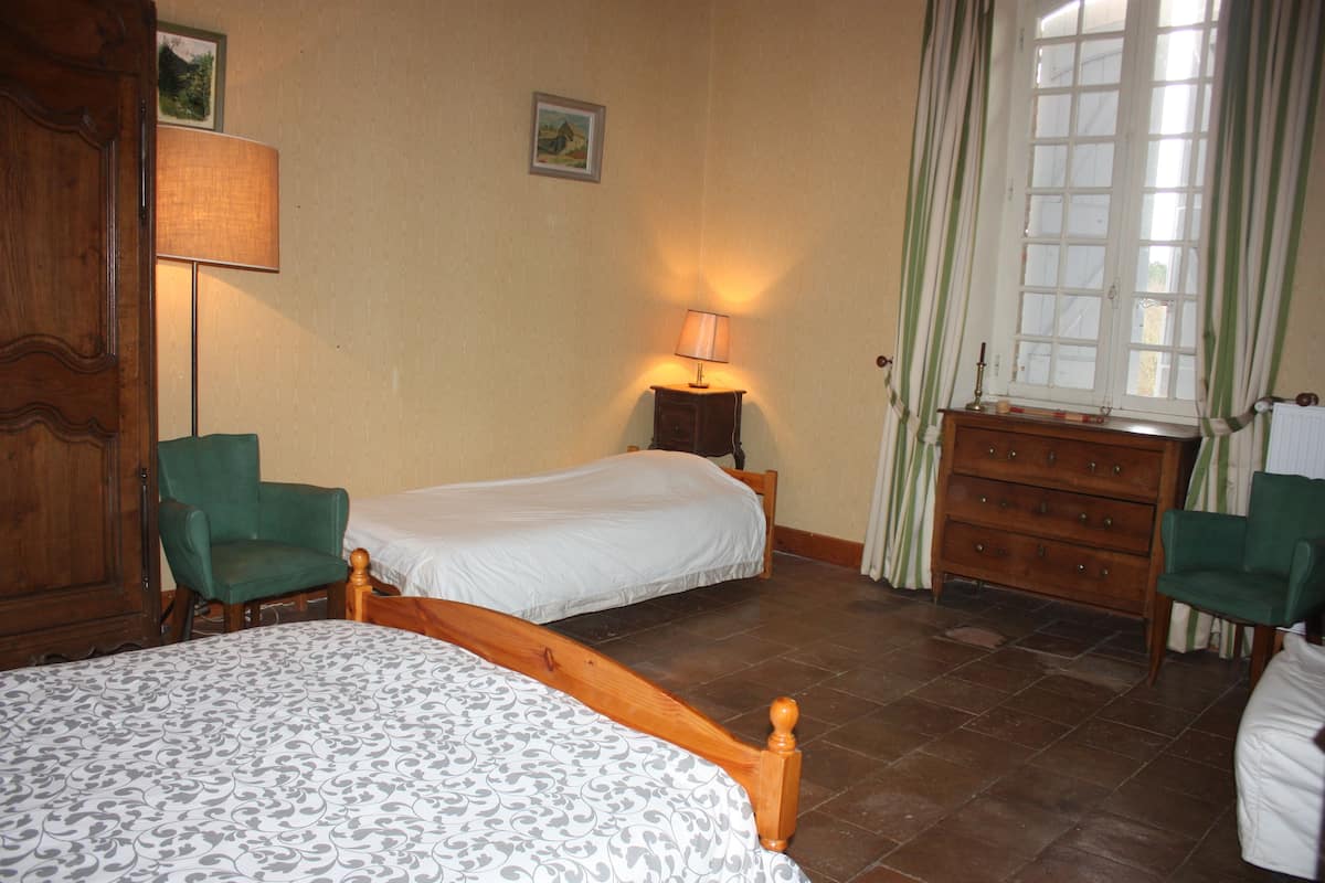 Une chambre avec deux lits simples, une commode en bois, deux chaises, une table de chevet avec une lampe et une fenêtre avec des rideaux.