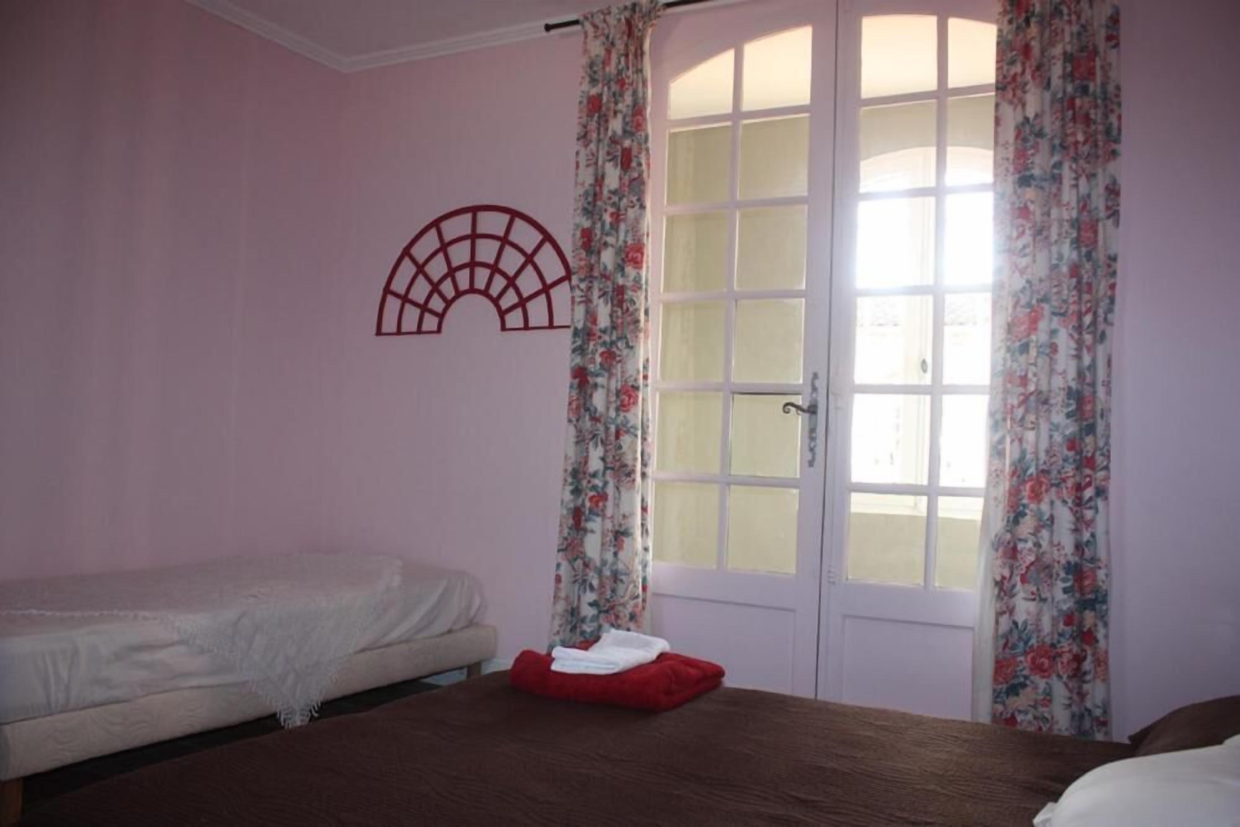 A bedroom with a bed, a window with floral curtains, and a door with a fan on the wall.