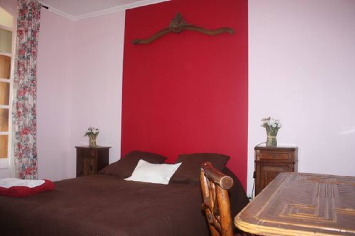 Une chambre à coucher avec un lit, une tête de lit en bois, un mur d’accent rouge, une petite table de chevet avec un vase et une fenêtre ornée de rideaux à motifs floraux.