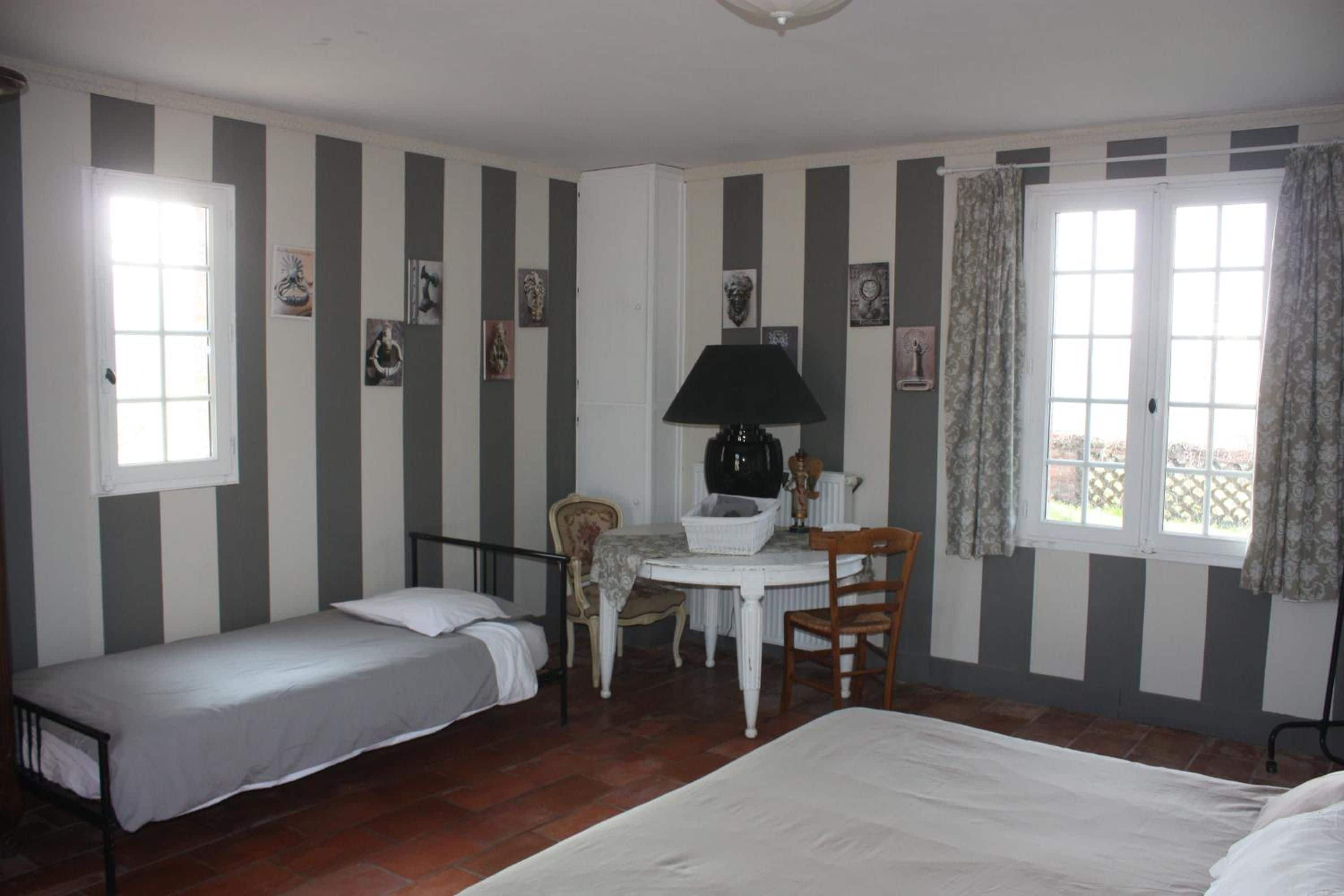 Ein Zimmer mit zwei Einzelbetten, einem kleinen Tisch mit einer Lampe und einem Sessel. Es gibt Fenster mit Vorhängen und Bilder an der Wand.