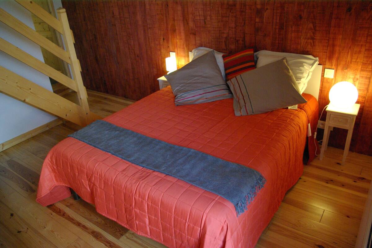 A bedroom with a wooden floor, a bed with an orange bedspread, pillows, and a bedside lamp.
