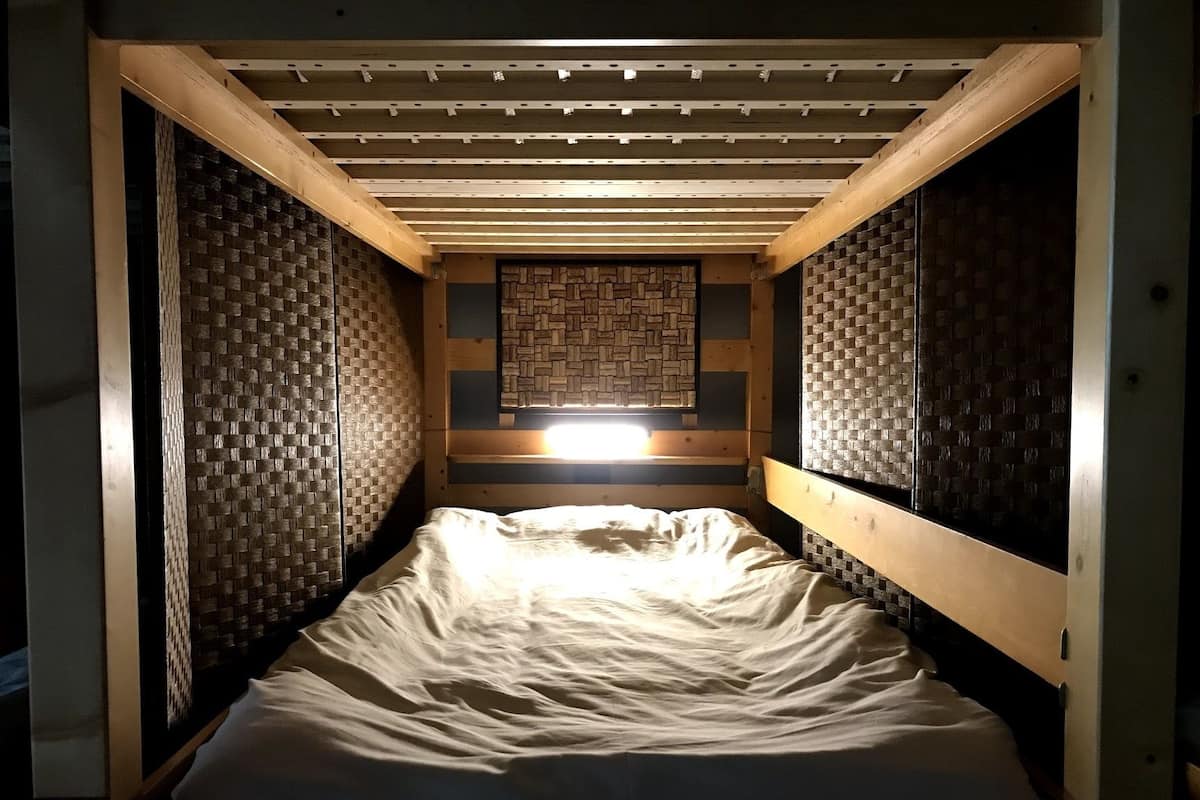 A narrow, single-bed room with a sloped ceiling and woven wall panels.