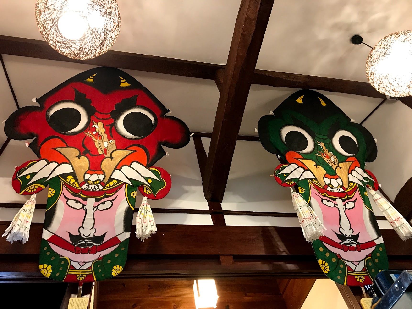 Two colorful masks hanging from the ceiling in a room with wooden beams and pendant lights.