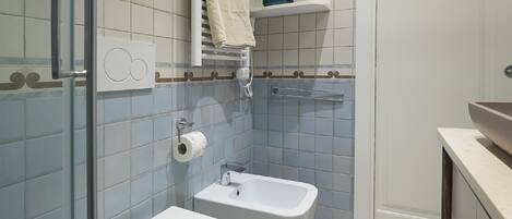 Un baño con dos lavamanos, una ducha e inodoro. Hay repisas sobre los lavamanos y una toalla colgando en la puerta de la ducha.