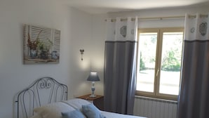 A bedroom with a bed, a nightstand with a lamp, a window with curtains, and a wall-mounted artwork. - Cante Grillet (Lacoste (Provence))