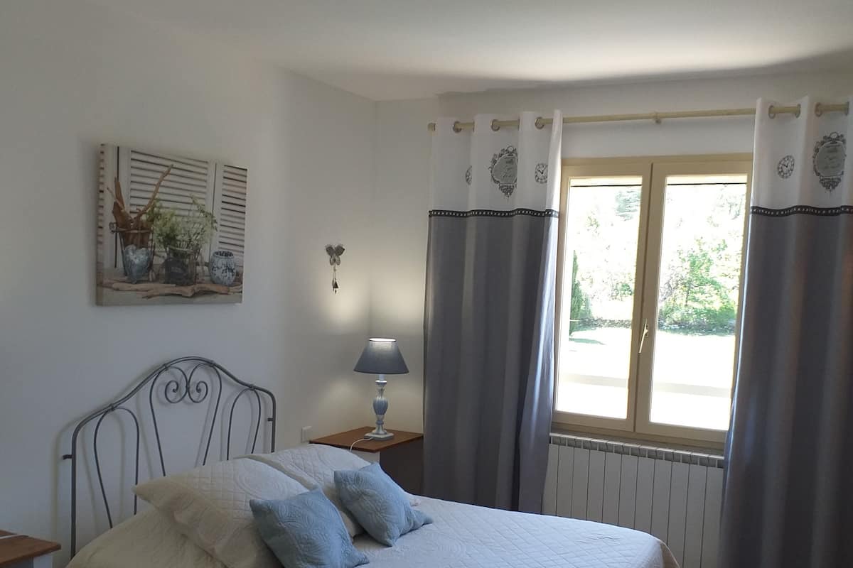 A bedroom with a bed, a nightstand with a lamp, a window with curtains, and a wall-mounted artwork.