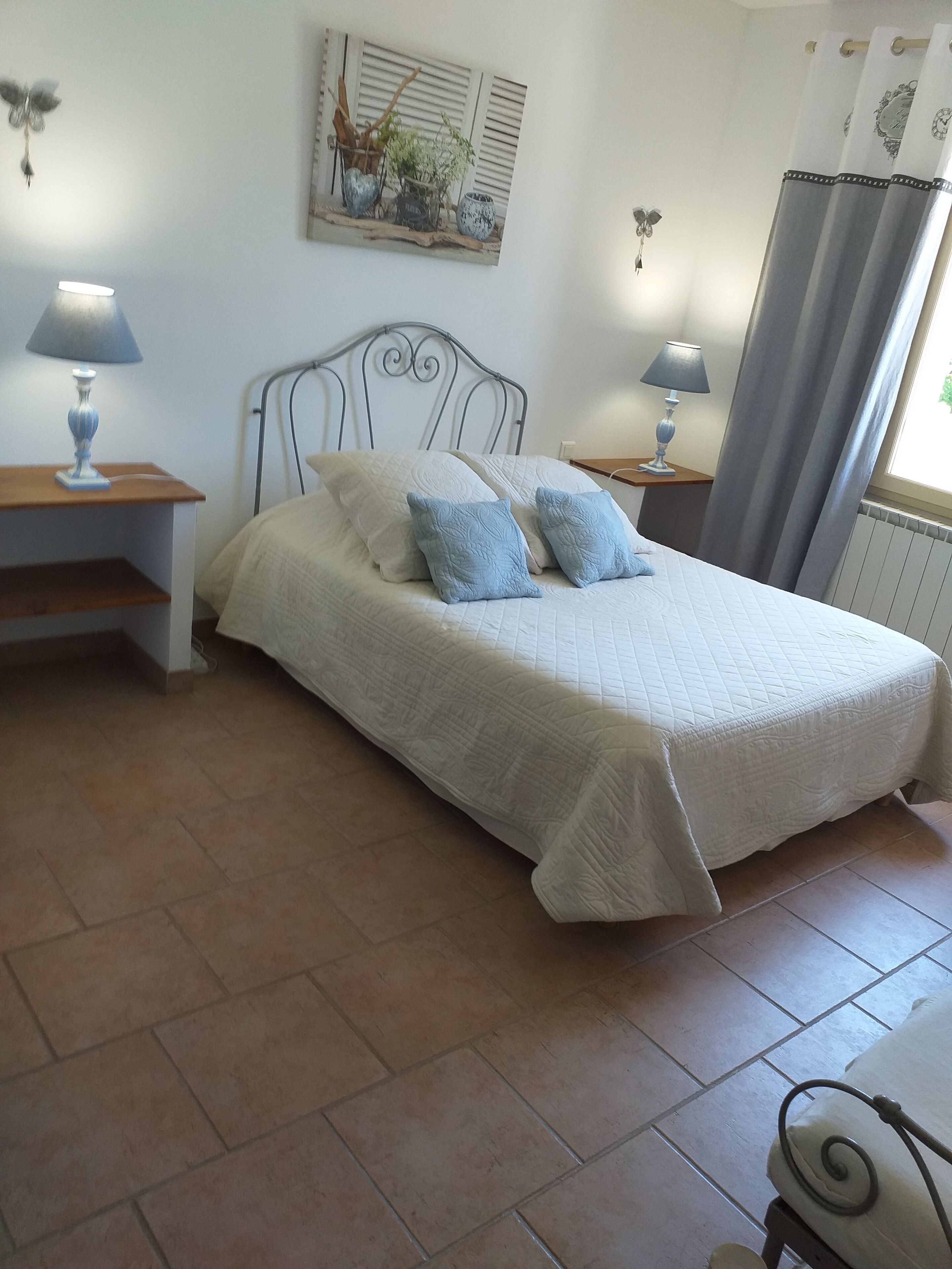 Un lit bien fait, recouvert d’une couette blanche et agrémenté de deux oreillers bleus, est flanqué de deux tables de chevet sur lesquelles reposent des lampes. Un tableau est accroché au mur, et une fenêtre est ornée de rideaux.