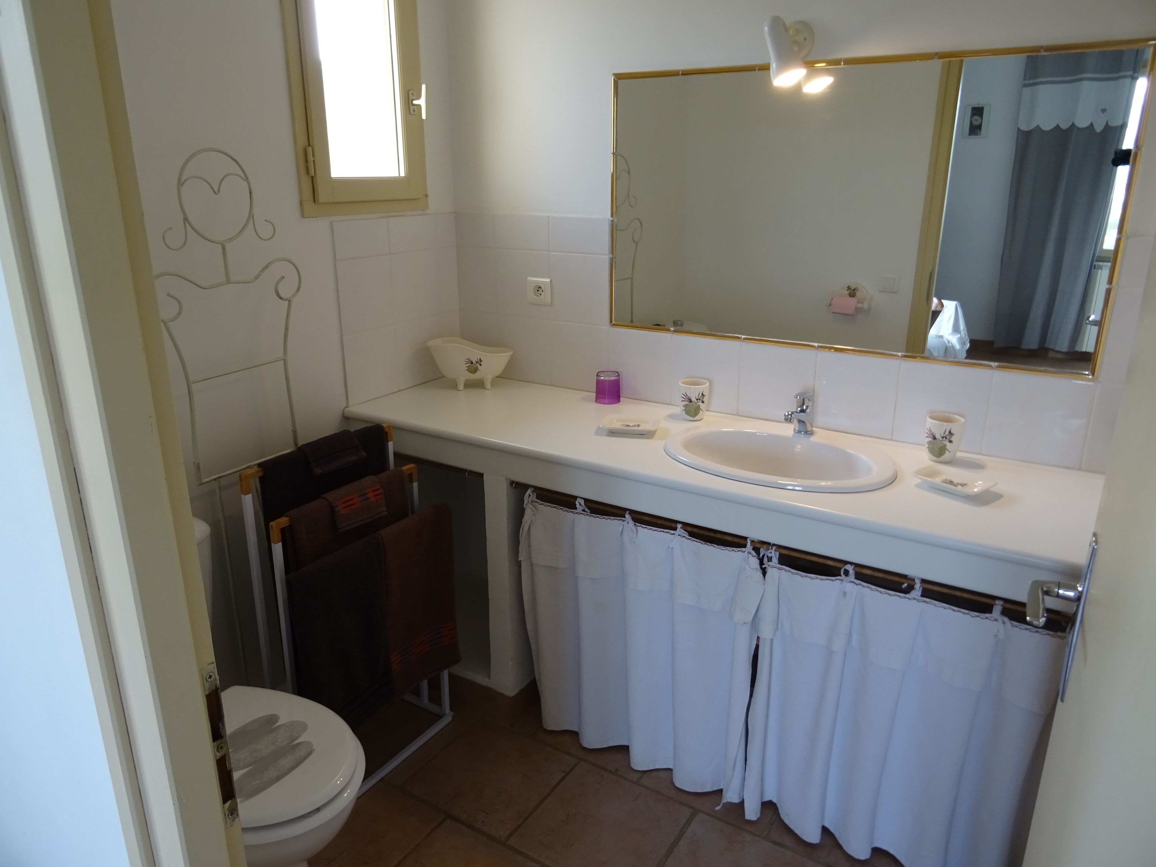 A bathroom with a toilet, sink, and mirror. There are towels hanging on a rack, a small shelf with toiletries, and a window with a view of the outside.