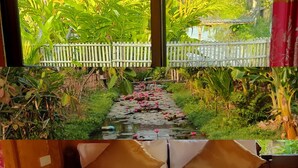Een bed met witte lakens en twee bruine kussens, een houten hoofdbord, en uitzicht op een tuin met een pad en waterlelies door een raam.