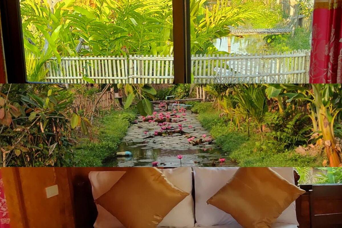 Un lit avec du linge de lit blanc et deux oreillers marron, une tête de lit en bois et, par la fenêtre, une vue sur un jardin avec un sentier et des nénuphars.