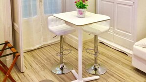 Una habitación pequeña con una mesa blanca y dos sillas blancas, un armario blanco y una ventana con cortinas translúcidas.