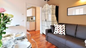 A modern living room with a grey sofa, a dining table set with plates and cutlery, a vase with flowers, and a kitchen in the background. - Vivaldi room - Biennale by Wonderful Italy (Venice)