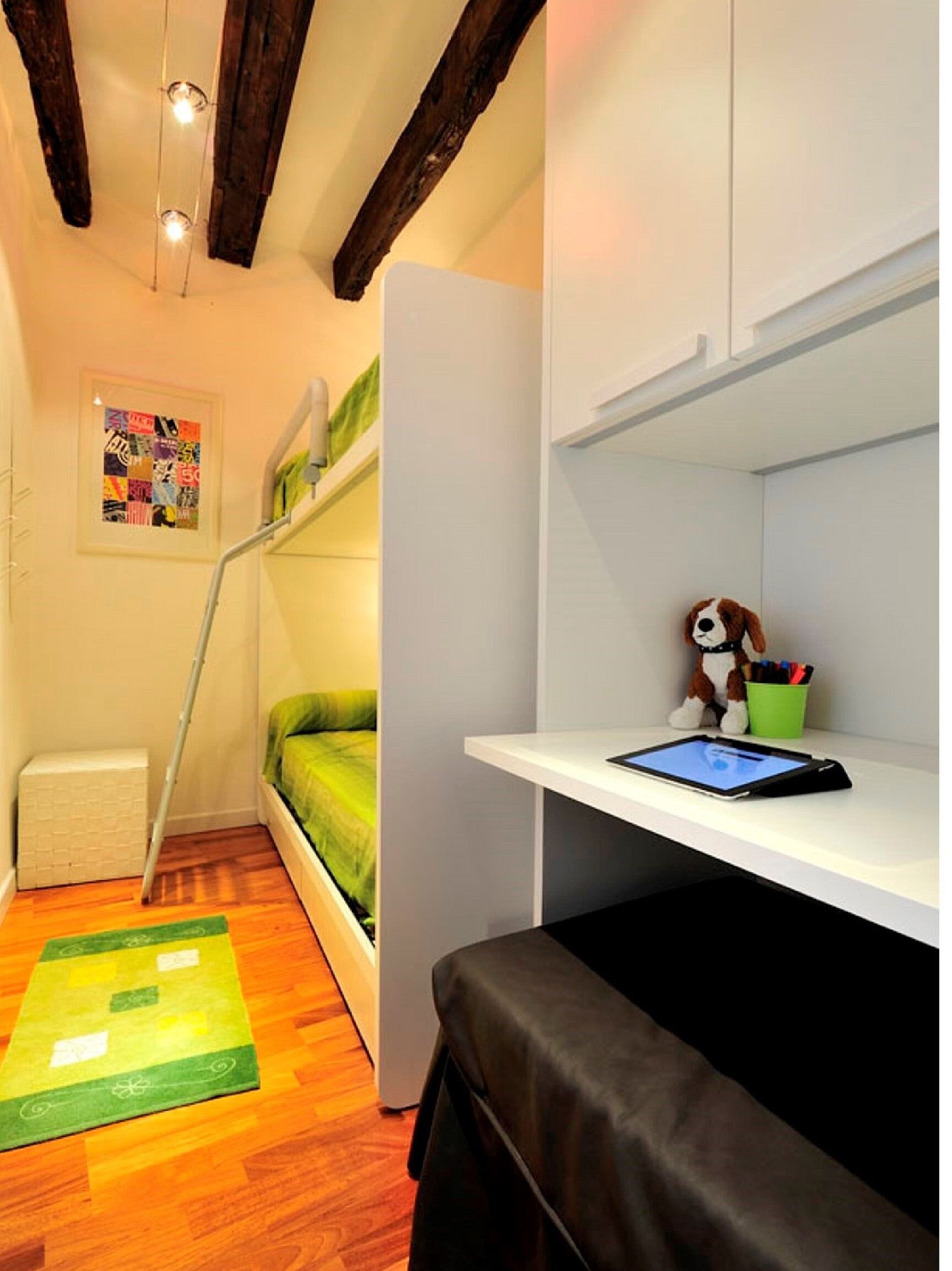 Ein kleines Zimmer mit einem Etagenbett, einem Schreibtisch mit einem Tablet, einem Stofftier und einem farbenfrohen Teppich.