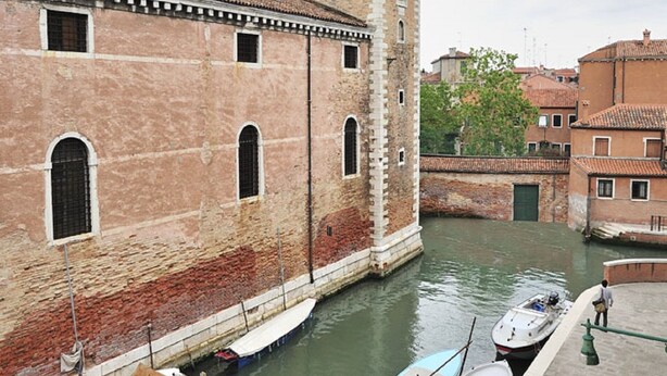 A canal with boats docked, a historic building with a tower, and a bridge in the background. - Vivaldi room - Biennale by Wonderful Italy (Venice)