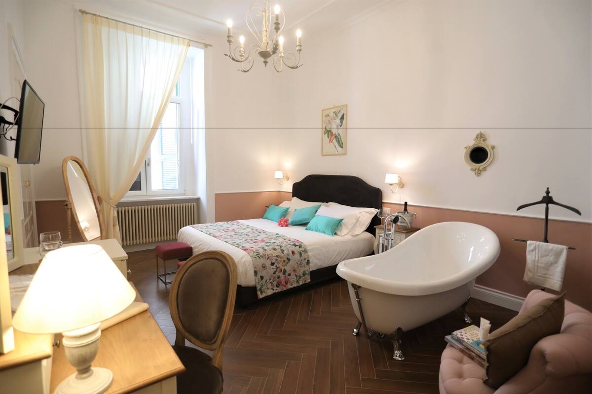 A bedroom with a bed, a vanity with a mirror, a lamp, a chair, a bathtub, and a desk.
