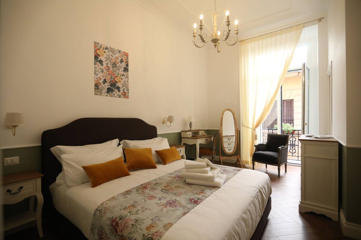 A neatly arranged bedroom with a large bed, a bedside table, a mirror, and a chair.