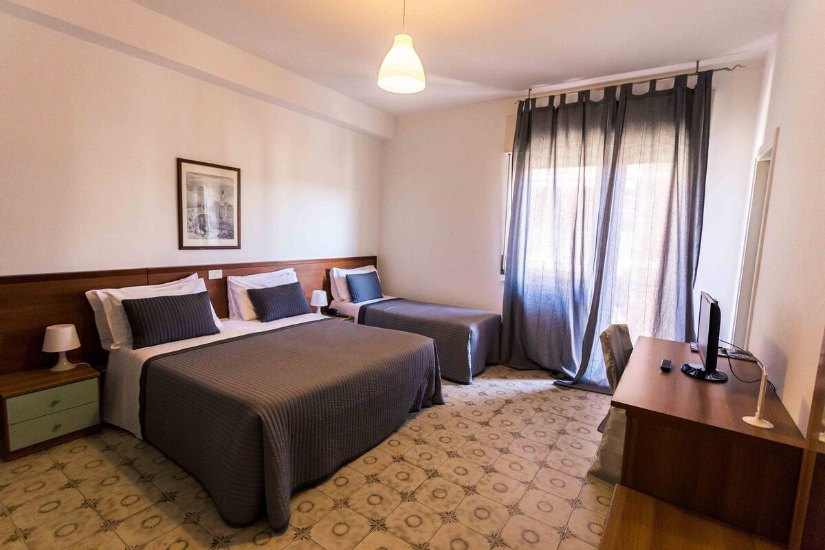 Une chambre d’hôtel avec deux lits, un bureau, une chaise, une lampe, un tableau au mur et une fenêtre avec des rideaux.