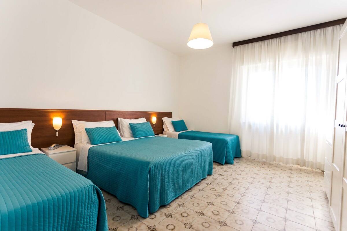 Une chambre d’hôtel avec une tête de lit en bois, deux lits avec une literie de couleur turquoise, une table de chevet avec une lampe et un sol à motifs.