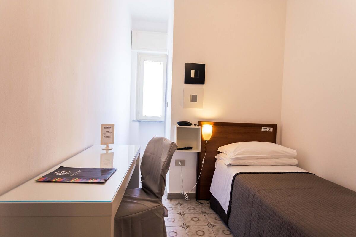 Une chambre d’hôtel comprenant un lit, un bureau avec une chaise, une table de chevet, une lampe et une fenêtre.