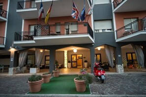 Entrada de hotel con alfombra verde, plantas en macetas, un scooter rojo y banderas.