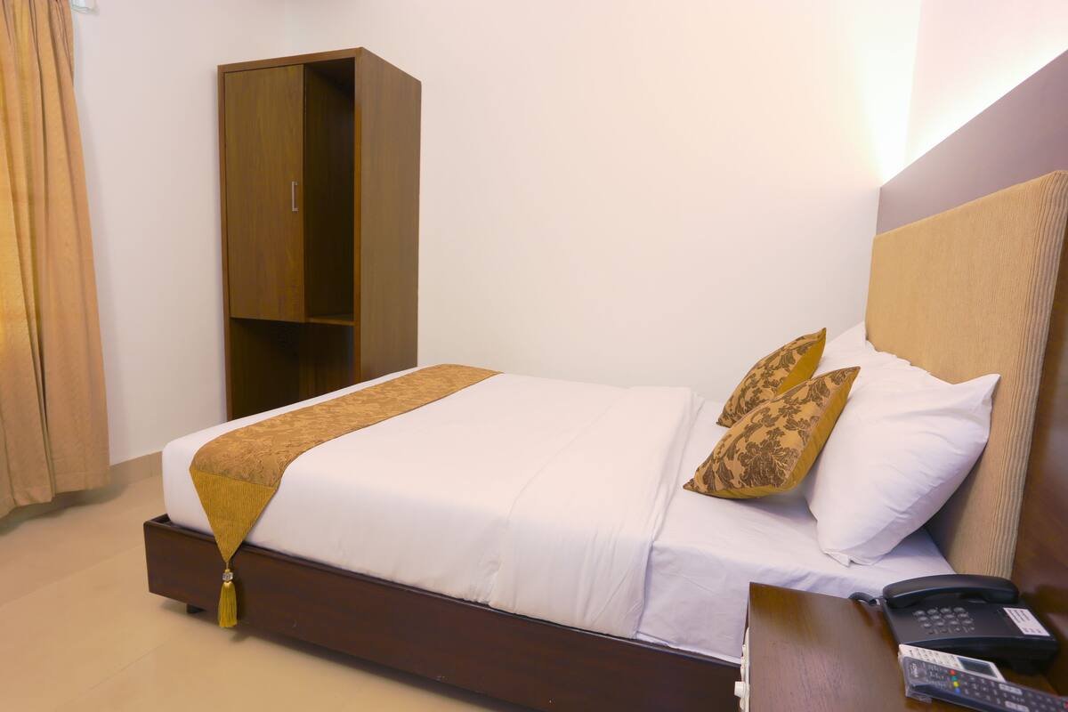 Ein ordentlich bezogenes Bett mit braunem Kopfteil, zwei Zierkissen, ein Nachttischchen mit Telefon und ein Kleiderschrank in einem Hotelzimmer.