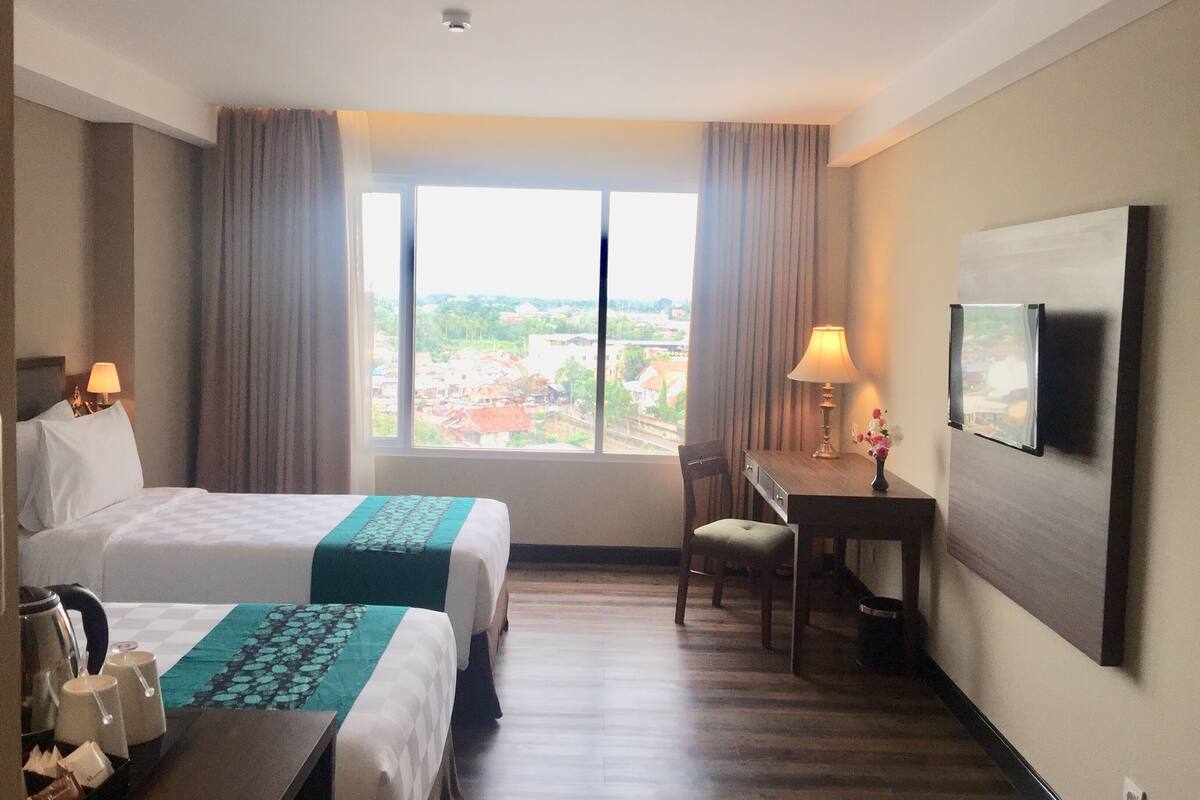 A hotel room with two beds, a desk, a chair, a TV, and a window with a city view.