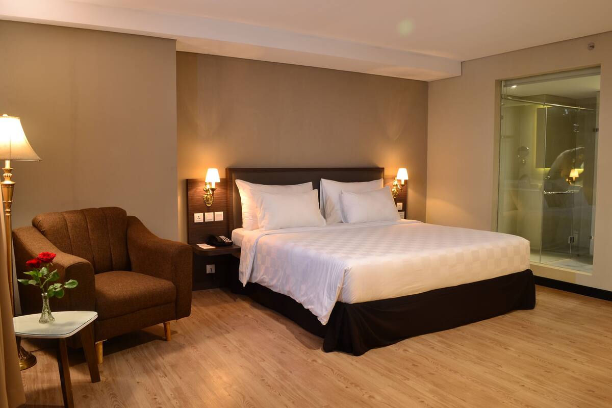 A hotel room with a large bed, a brown armchair, a small table with a vase of flowers, and a glass shower enclosure.