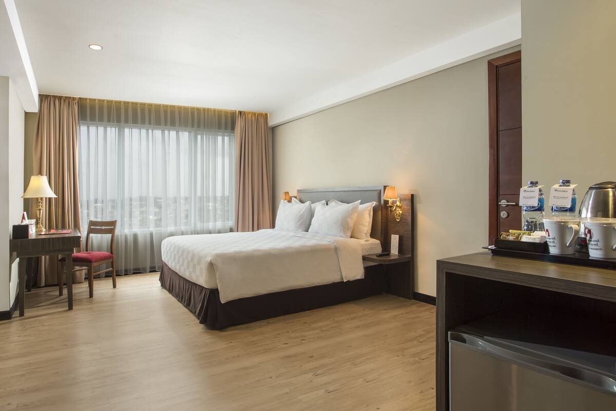 A hotel room with a bed, bedside table, lamp, TV, desk, chair, and a minibar.