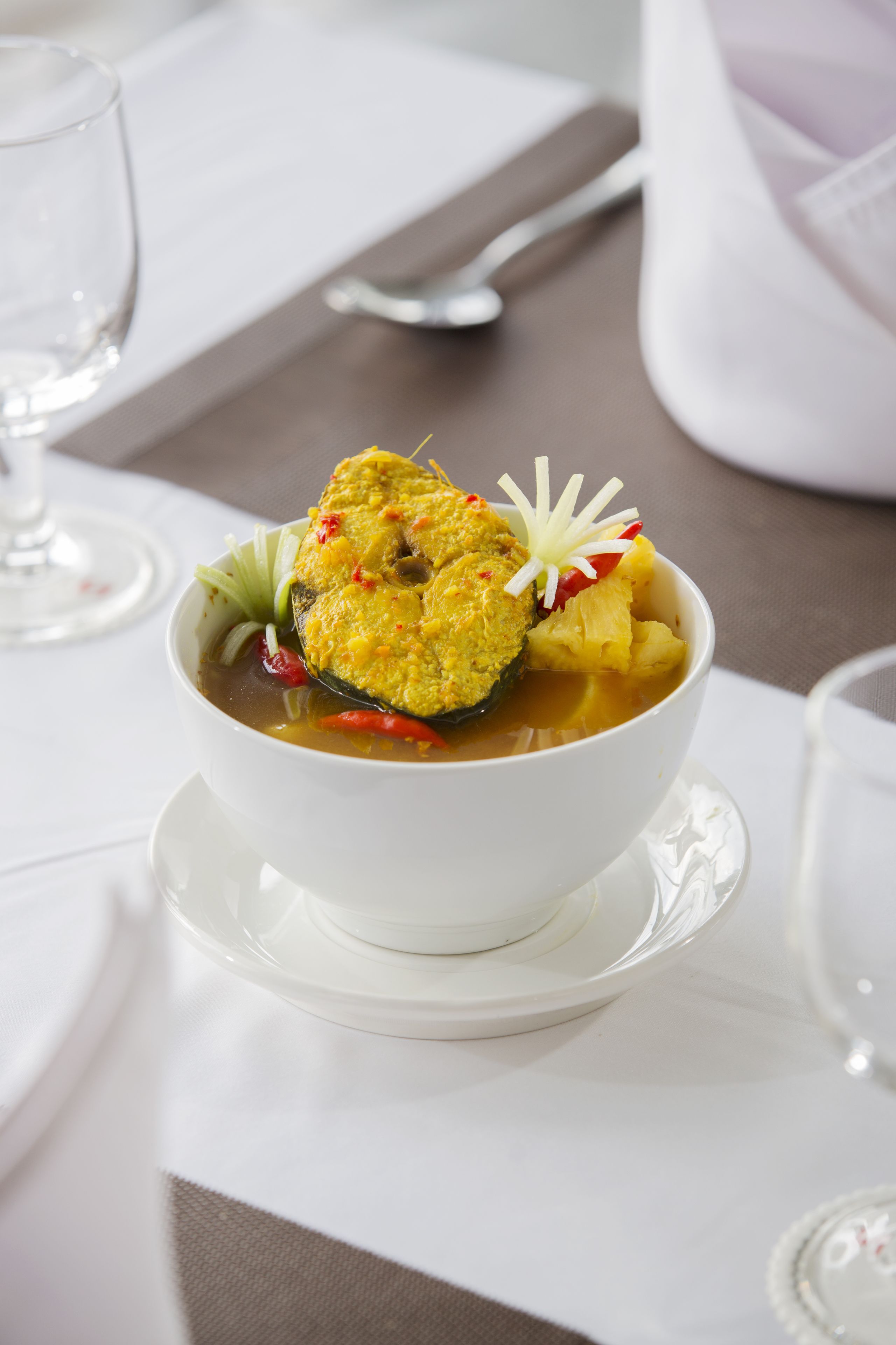 Un tazón blanco de sopa con decoración de pescado sobre un plato blanco, acompañado de una cuchara y un vaso de agua sobre una mesa.