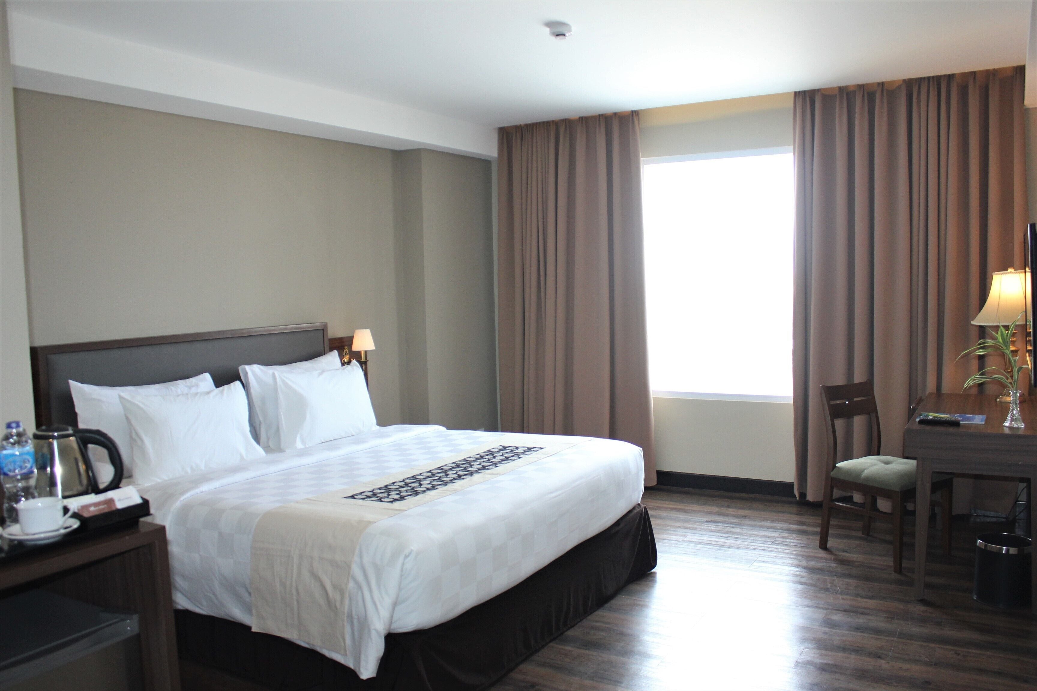 Une chambre d’hôtel avec un grand lit, un bureau avec une chaise, une machine à café et une fenêtre avec des rideaux.