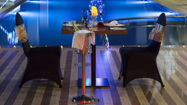 Un área de comedor junto a la piscina, con una mesa dispuesta para dos personas, rodeada de velas, y una piscina al fondo.